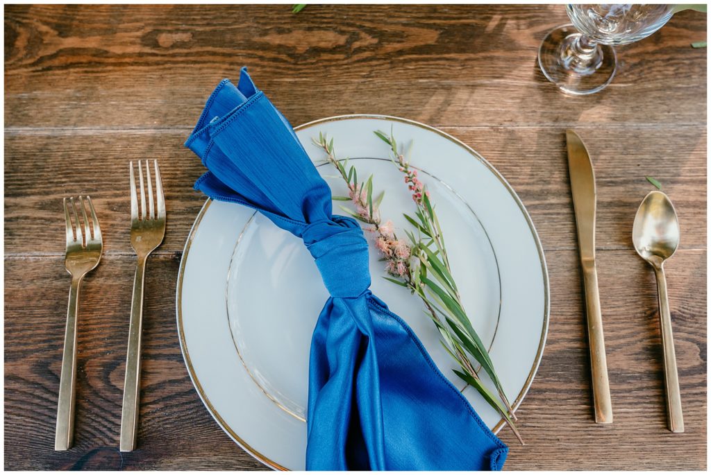 wedding place setting with greenery and gold silverware with navy blue napkin
