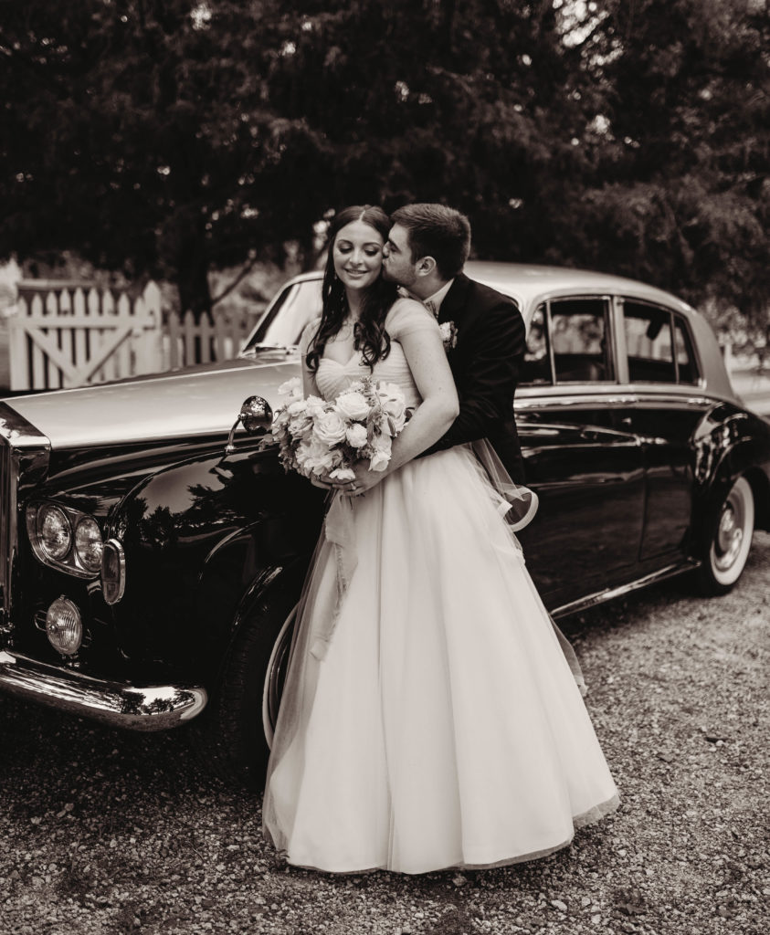 Bride and groom hugging in front of old car after wedding