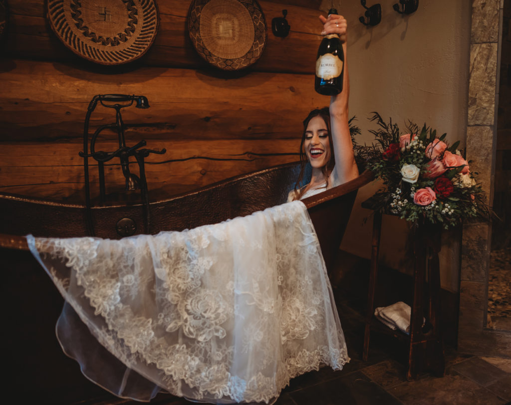 bride sitting in bath tub in her wedding dress holding bottle of champagne celebrating her wedding day