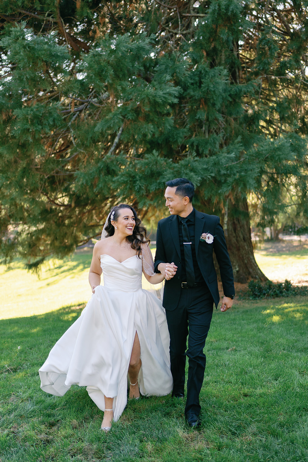Newlyweds walk through a park under a tree holding hands and smiling at each other