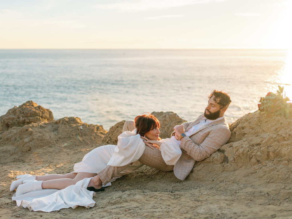 A bride lays in the lap of her groom in a plaid suit on a rocky oceanside trail at sunset thanks to San Francisco Wedding Planners