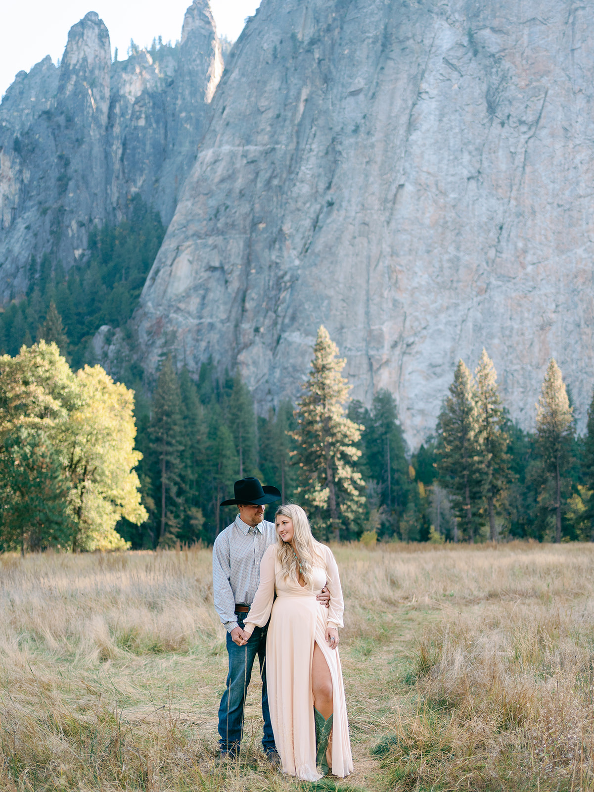 Newlyweds in a pink dress and a a cowboy hat stand holding hands in a field of tall golden grass in a mountain valley