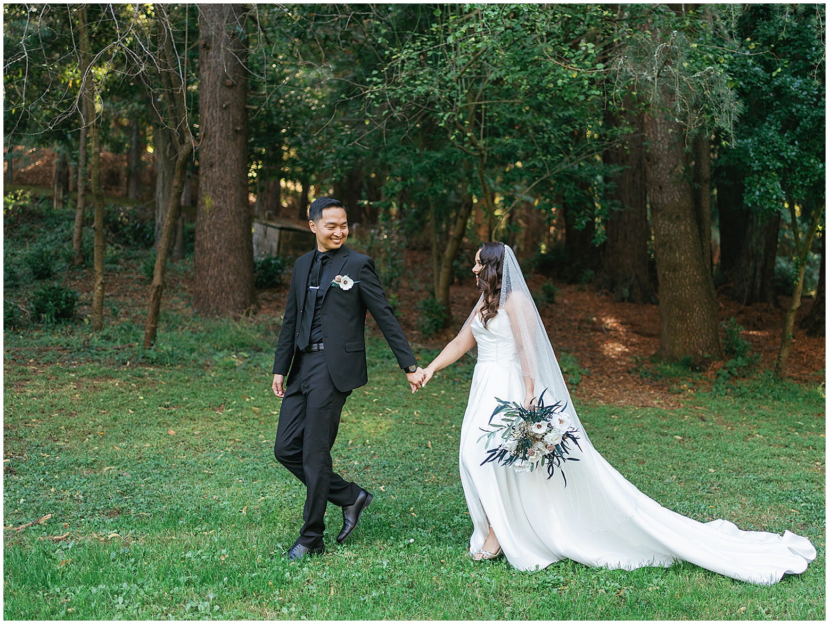 A groom in a full black suit leads his bride by the hand through a forest lawn