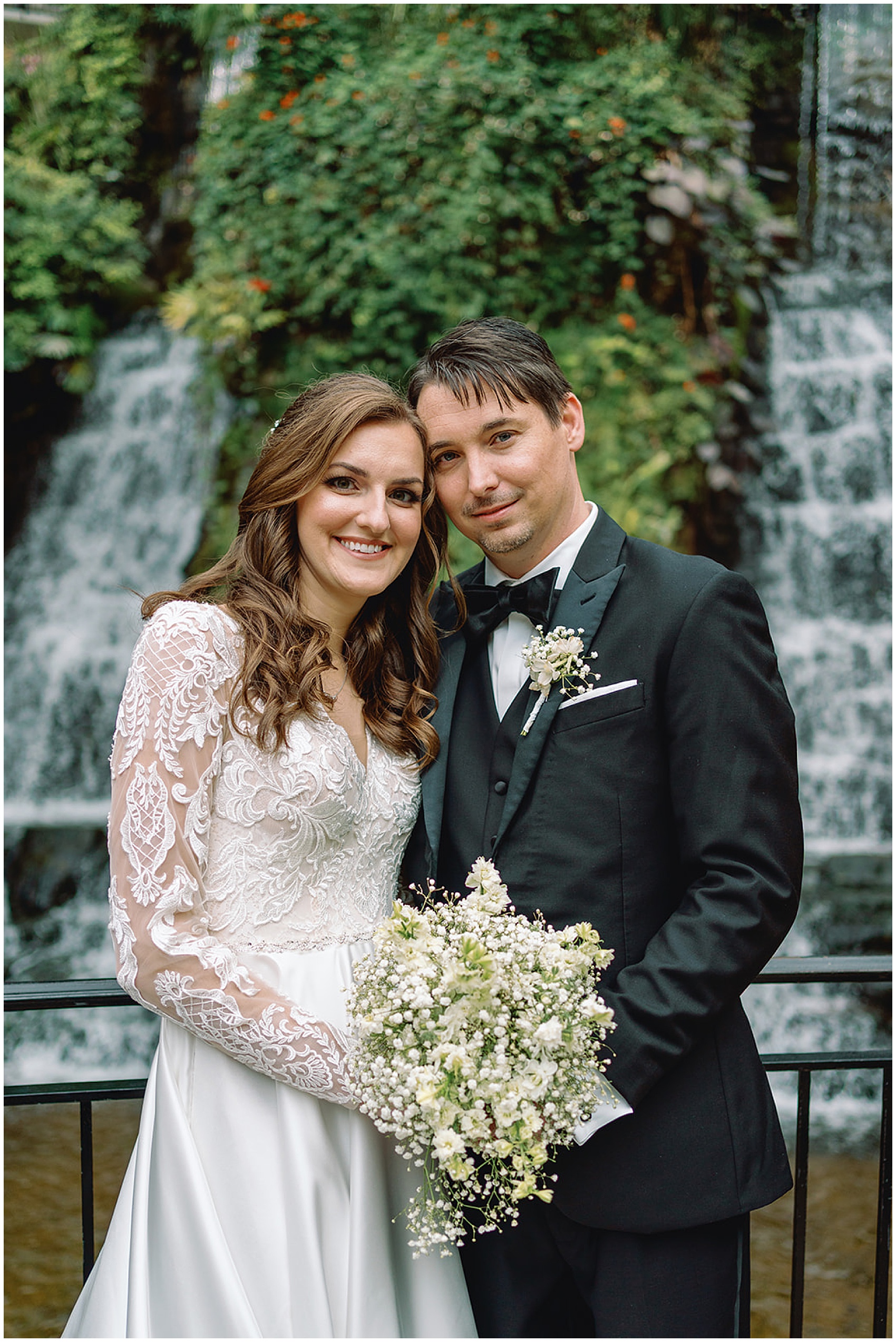 Newlyweds stand together in front of a large waterfall holding the white bouquet
