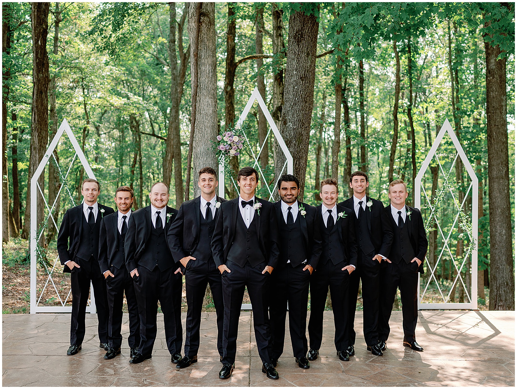A groom in a black tuxedo stands with hands in his pockets with his matching groomsmen
