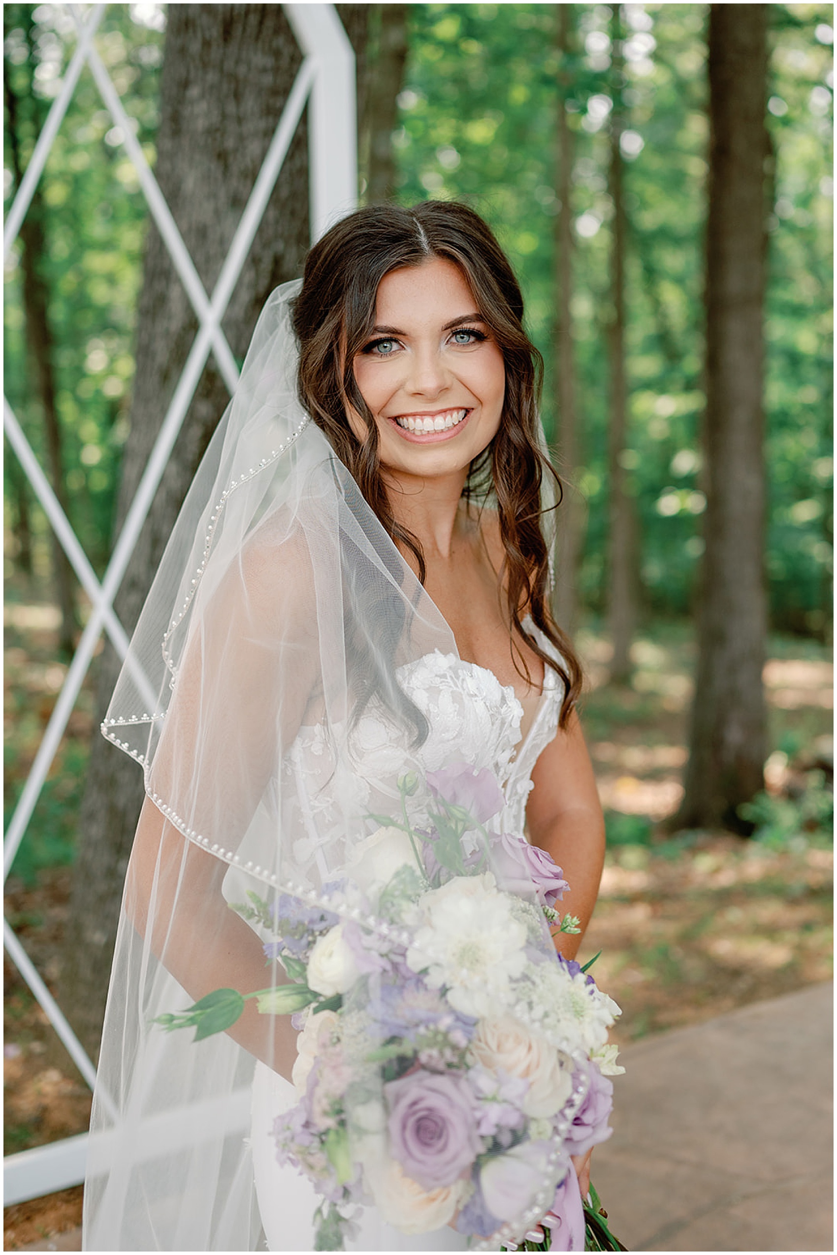 A bride smiles while standing in a forest holding her purple and white bouquet at her hickory meadow charlotte tn wedding