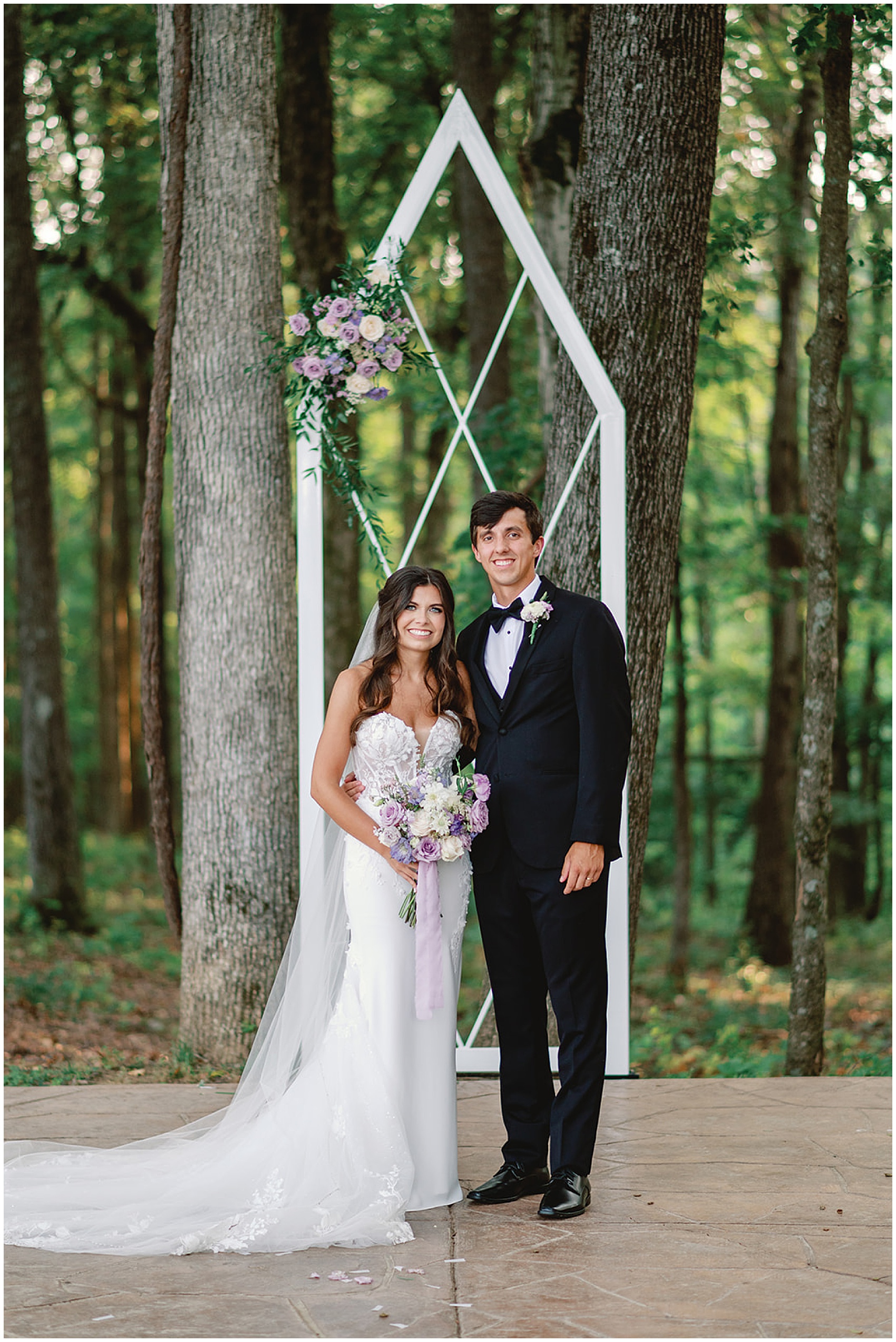 Newlyweds smile and stand together on a forest patio