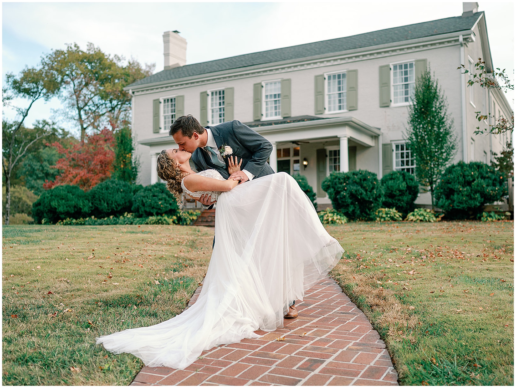 A groom dips and kisses his bride while standing on the brick front path to the maple grove estate