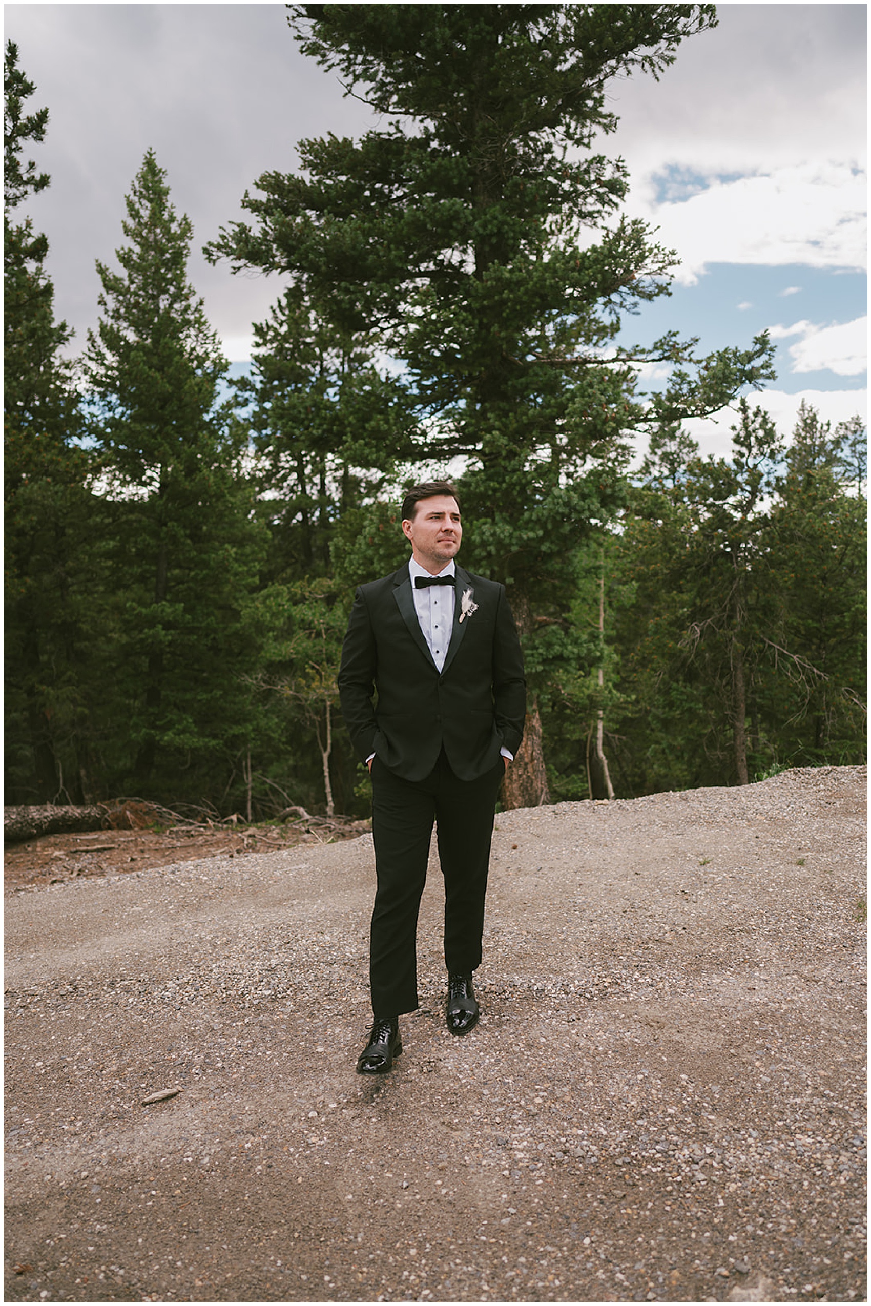 A groom in a black tuxedo stands on a gravel road with hands in pockets