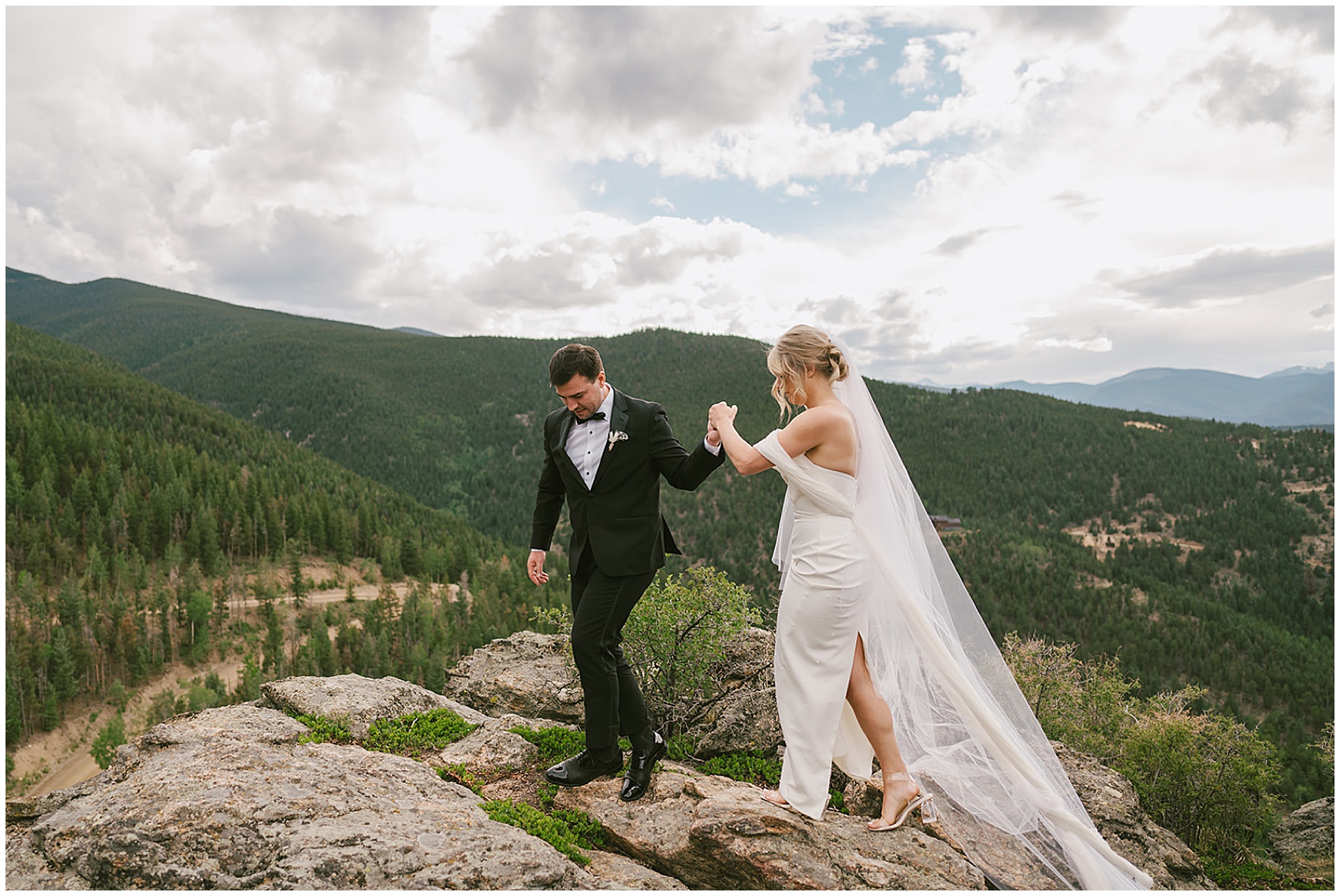 A groom in a black tuxedo leads his bride up a mountain rock overlook