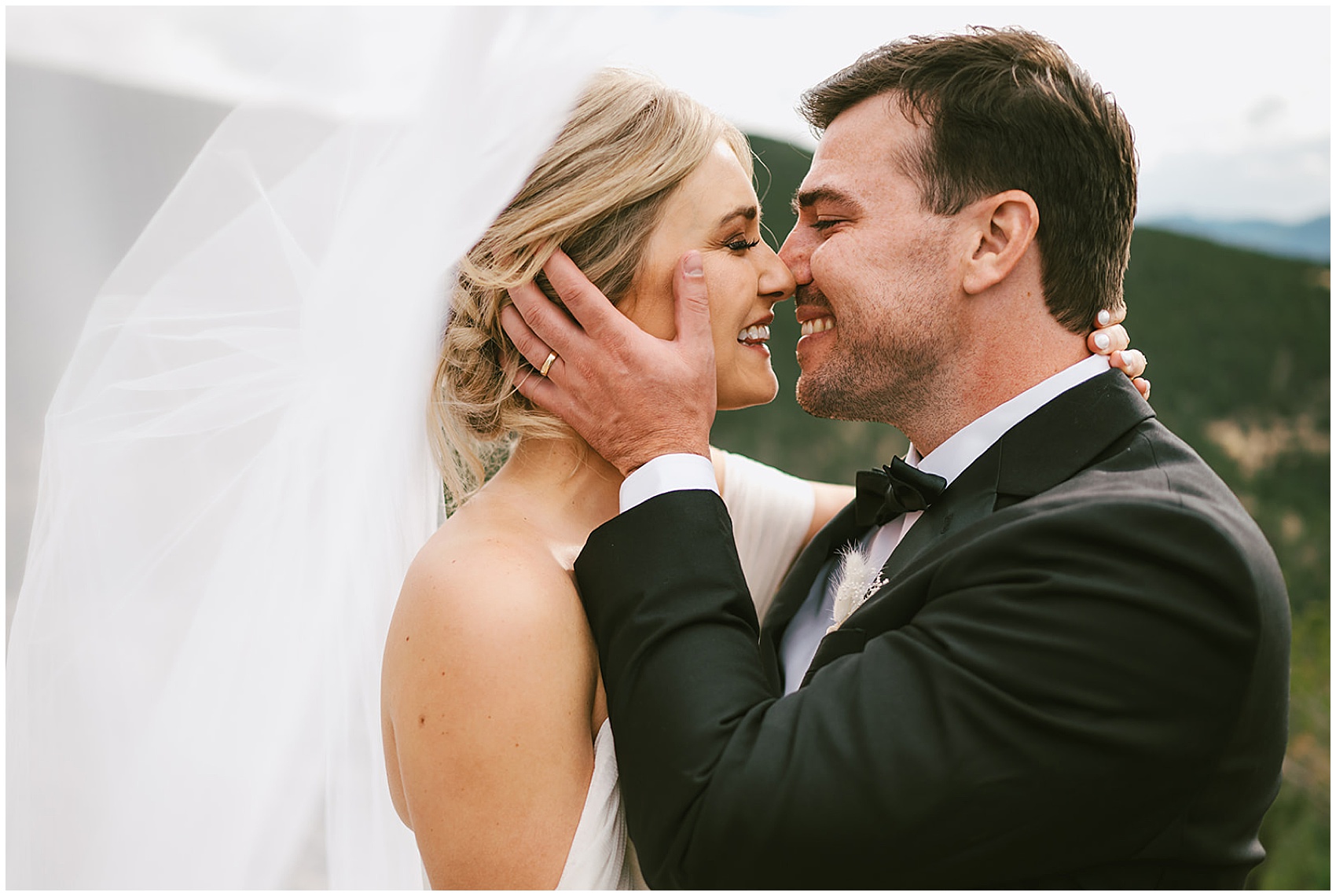 Newlyweds lean in for a kiss in a black tuxedo and white dress with a long veil flowing in the wind