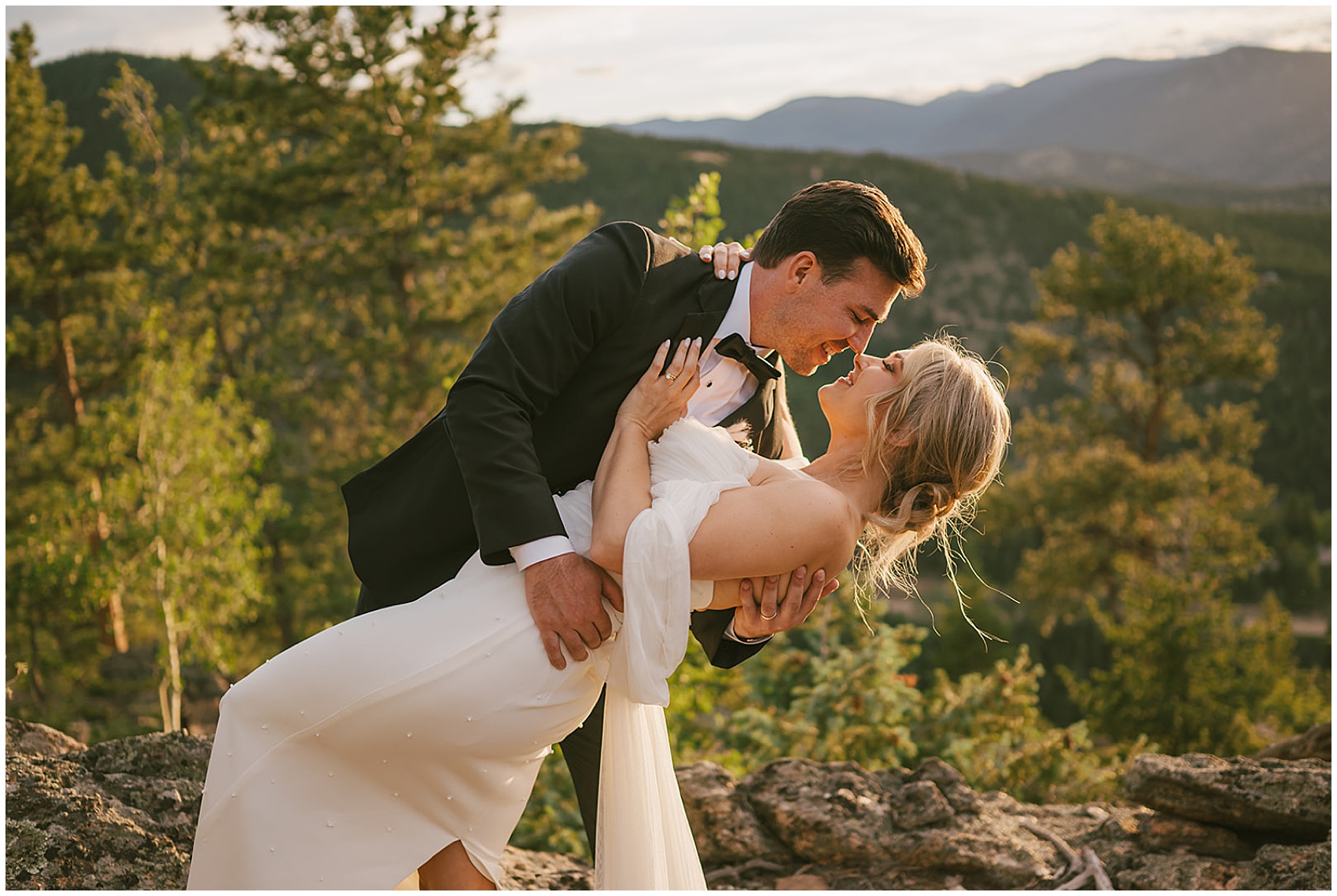 A groom in a black tuxedo dips and kisses his bride on a mountain trail at their north star gatherings wedding