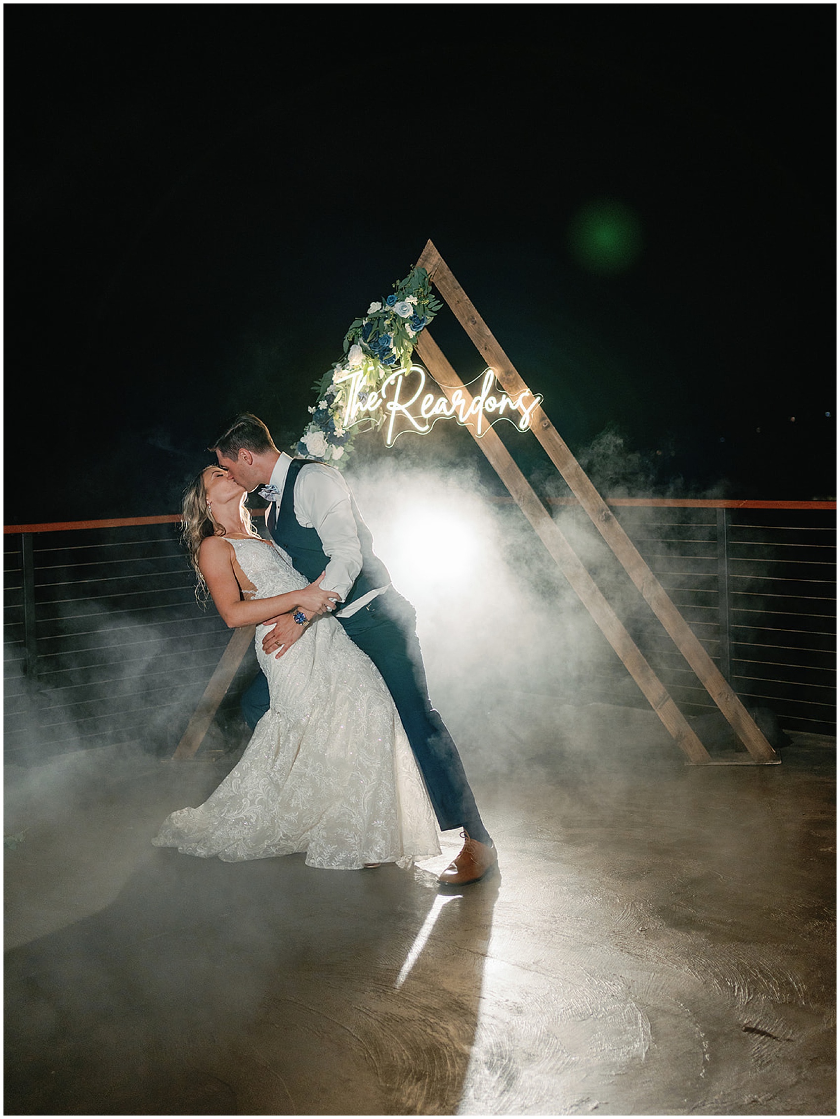 A groom dips and kisses his bride in front of a custom wood triangle arbor with a smoke machine