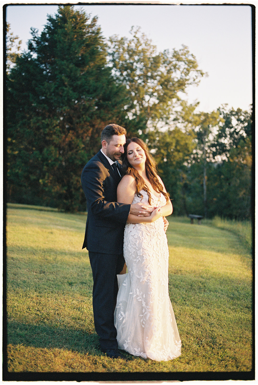 A groom hugs his bride from behind as she leans onto him in a lawn at sunset at their Firefly Lane Chapel wedding