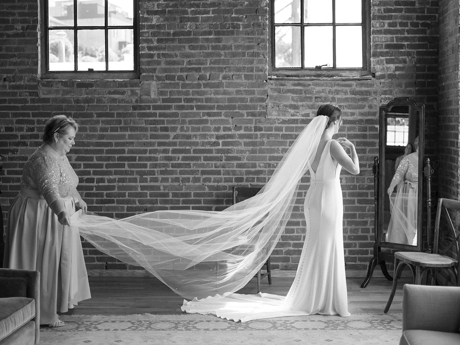 A bride stands in a mirror getting ready in a brick room while mom spreads her veil at The Standard Wedding Venue